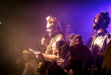 A drag performer in gold sequins sings into a microphone during a vibrant live theater production.