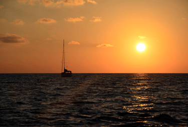 Atardecer, con el cielo anaranjao y un velero de fondo, con las velas recogidas, en medio del mar