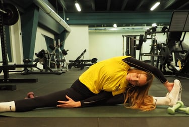 Una donna su un tappetino da yoga in palestra esegue una posa laterale di stretching, tenendo il piede con una mano mentre si