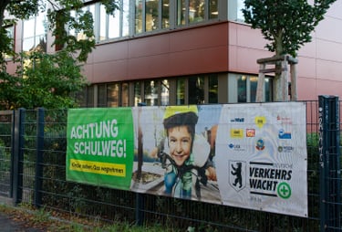 Foto von einerm roten Schulgebäude, an dem Zaun davor ist ein Plakat zur Schulwegsicherheit.