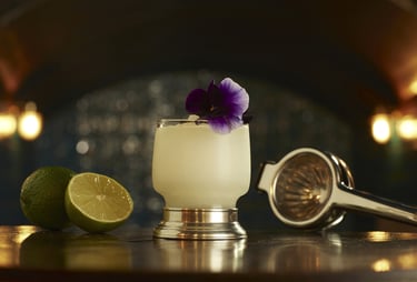 White cocktail with a pansy flower garnish next to cut limes and squeezer in an art deco bar