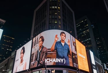 A wide shot of a modern city at night with digital screens displaying vibrant advertisements. Dark navy and tan color palette. International setting.