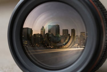 Extreme close-up of a camera lens reflecting a North American / US city skyline at twilight, muted champagne gold light trails, deep obsidian black glass.