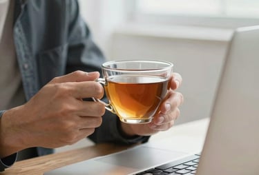 A close-up of a person's hands holding a cup of tea while looking at a laptop in a bright room. The atmosphere is calm, collaborative, and professional.