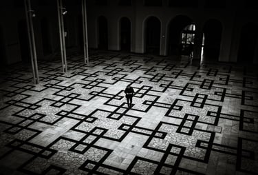 High-contrast black and white photograph of a lone figure framed by dramatic light on a large, patterned stone floor.