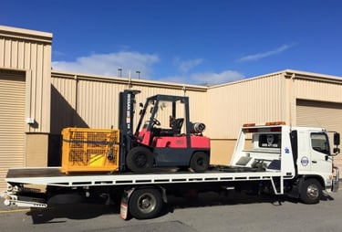 A Nissan forklift and yellow storage cage loaded onto a white tilt tray towing truck for transport.