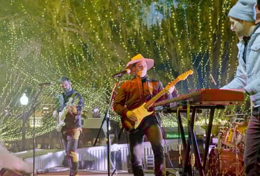 Musician performing live on stage under bright decorative string lights, playing electric guitar bes