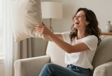A warm, candid photo of a stylish mom laughing while sitting on a chic neutral armchair, playfully throwing a cream-colored throw pillow. Bright, funny, and beautiful atmosphere in a North American / US living room.