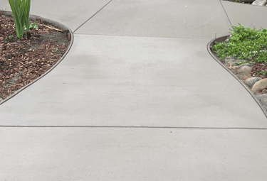 Curved concrete walkway in a backyard garden with landscaping, ferns, and patio furniture.