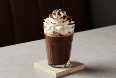 A professional food photography shot of an artisanal dark chocolate mocha topped with whipped cream and cocoa shavings, set on a beige stone coaster in a minimalist North American / US cafe.