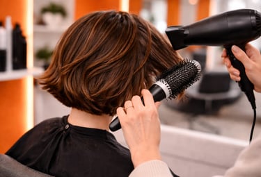 Professional hairstylist using a round brush and blow dryer to style a brunette bob haircut in a salon.