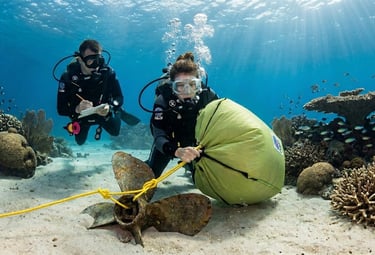 Scuba divers using a yellow lift bag to recover a sunken boat propeller from a coral reef seabed.