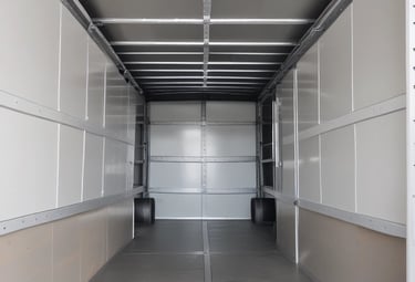 A black truck parked in an industrial area is loaded with large metal cabinets and a roll of white material. The truck's bed is wooden and surrounded by various parked vehicles in a warehouse environment.