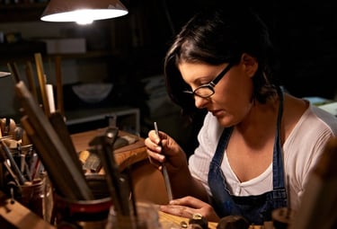Female jeweler using hand tools to craft custom jewelry at a wooden workbench.