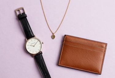 Elegant flat lay of fashion accessories including a minimalist watch, a delicate gold necklace, and a high-quality leather cardholder on a light lilac background. Soft, feminine lighting, South American / Brazilian fashion boutique style.