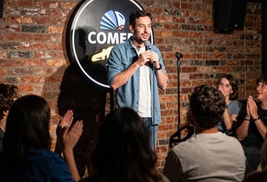 Stand-up comedian performing a live comedy set on stage for a laughing audience in a brick-walled club.