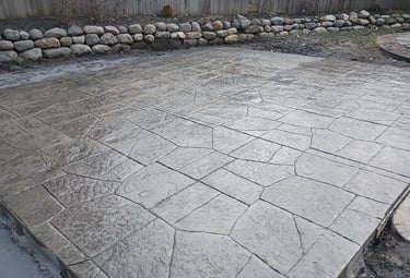 Freshly poured gray stamped concrete patio with flagstone pattern and rock wall landscape border.