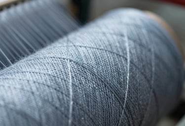 A macro photograph focusing on the intricate texture of Kota yarn. The thread is crisp and well-defined against a soft-focus background of the spinning machinery. Lighting highlights the metallic sheen of the fiber, incorporating #95A5BB tones.