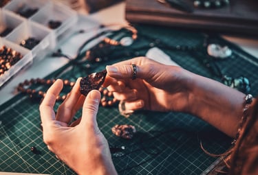Artesana sosteniendo una piedra preciosa sobre fondo verde con otras piedras y materiales 