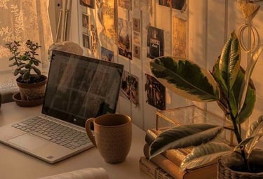 esquina de un escritorio con plantas, una taza y una computadora con los rayos del sol dorados entrando por la ventana