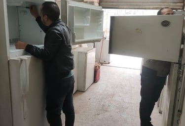 Professional technician in a black jacket repairing a white refrigerator in an appliance service shop.