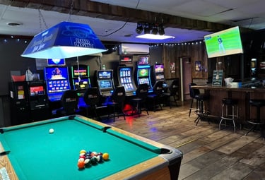 Rustic sports bar interior with a green pool table, neon slot machines, and a wooden bar area.