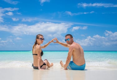 A couple making a heart shape with their hands on a tropical beach with turquoise water.