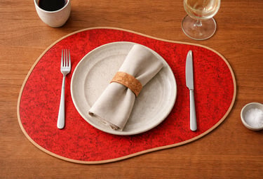 Red oval placemat with a ceramic dinner plate, linen napkin, and silverware on a wooden table.