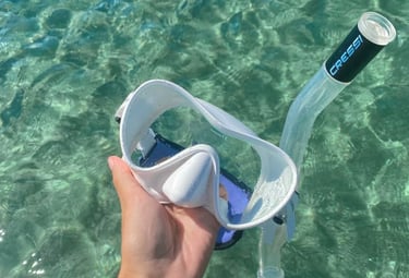 A hand holding a white Cressi snorkeling mask and snorkel over clear turquoise ocean water.