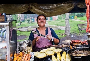 A smiling street food vendor grilling corn on the cob, sausages, and arepas at an outdoor market.