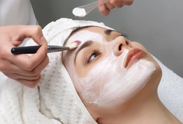 A woman with a white towel on her head receiving a professional facial treatment with a  brush