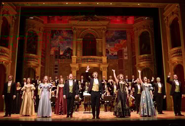 A wide, dramatic shot of a grand opera stage during a live performance in a North American opera house. The stage is illuminated with rich, warm golden and deep red theatrical lighting, highlighting elaborate, classical scenery that suggests an opulent historical setting. Soloists and chorus members in period costumes are performing a climactic scene, their faces conveying intense emotion and their voices projected with powerful expressions. An orchestra is visible in the pit below, led by a conductor with an impassioned gesture. The composition captures the spectacle, artistic excellence, and emotional depth of a live operatic production, with a sophisticated and timeless aesthetic.