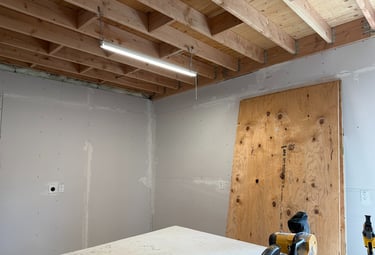 Basement renovation progress with exposed ceiling joists, drywall installation, and shop lighting.
