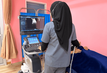 A healthcare professional in a hijab performing a diagnostic ultrasound on a patient in a pink medical room.