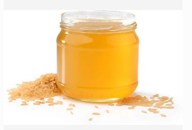 A glass jar of yellow rice bran oil surrounded by scattered grains of brown rice on a white background.