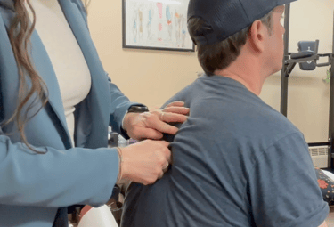 Chiropractor pinching seated patients back during exam with treatment room in background.