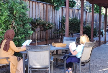 Two women at a table outside reading. One with a hijab, one with light hair.
