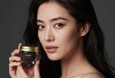A captivating portrait of an elegant North American / Western European woman, late 20s, with impeccably radiant, dark, flowing hair, gently holding a sleek, dark glass HARIR Caviar vial. She gazes confidently with soft, professional makeup that enhances her natural beauty. The composition is a sophisticated, close-up lifestyle shot, emphasizing her elegant hands and glowing complexion. Dramatic, soft studio lighting creates a luxurious, high-contrast effect, with subtle golden reflections catching the edge of the vial and her hair. The background is a blurred, deep charcoal grey, maintaining a dark, luxurious aesthetic. The overall style is modern, aspirational, and perfectly suited for a high-end beauty brand's Instagram.