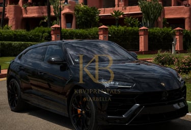 Sleek black Lamborghini Urus SUV parked in front of a luxurious peach-colored building with manicured landscaping, Marbella