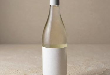 A studio-lit photograph of an elegant white wine bottle on a sand-colored stone surface. The lighting is soft and directional, highlighting the texture of the artisanal paper label.