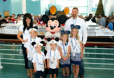 A family poses with Mickey Mouse in front of a Disney Cruise Line ship model at a port terminal.