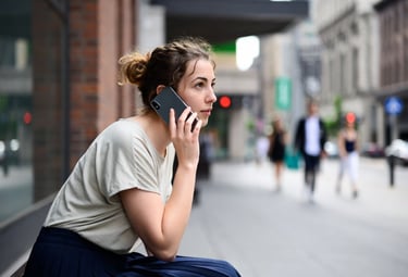 Young woman talking on a smartphone while sitting on a city street sidewalk.