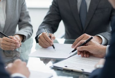 A close-up photograph of a professional meeting in a North American corporate setting. Two individuals in business attire are discussing documents on a clean glass table. The lighting is bright and clear.