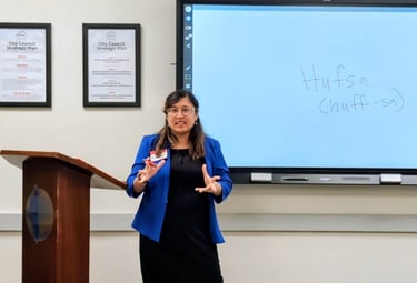 A woman in professional clothing with a blue blazer is speaking in front of a whiteboard, two awards, and a podium.