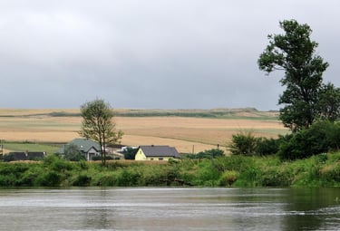 Widok ze śrdoka rzek blisko wody. Rzeka zakręca a na zakręcie w oddali domy. Dalej domy i pole