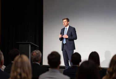 A professional man in business attire speaking from a stage in a North American conference hall. The focus is on the speaker, with a soft-focus audience in the foreground. Lighting is professional and highlights leadership and authority.