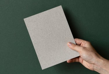 A professional shot of a designer’s hand holding a high-quality textured paper sample against a matte forest green wall.