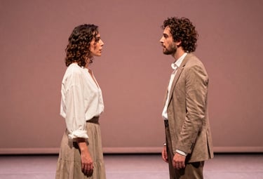 Two actors in an intense dialogue on a minimalist stage, wearing elegant Southern European attire, lit from the side with a warm, dusty rose hue.