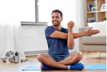 Man sitting cross‑legged while stretching his right arm across his chest, using his left arm to gently support the stretch.