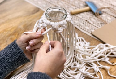 Manos haciendo nudos para una funda de macramé DIY para frasco, utilizando cordón de algodón blanco 